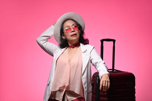 Confident middle age woman in sunglasses and straw hat standing with suitcase on pink background.