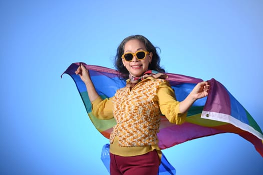 Older lady holding a rainbow flag on blue background LGBT, human rights, freedom and equality concept.