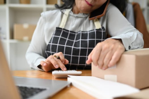 Middle age female small business owner having phone conversation and calculating expense at desk.