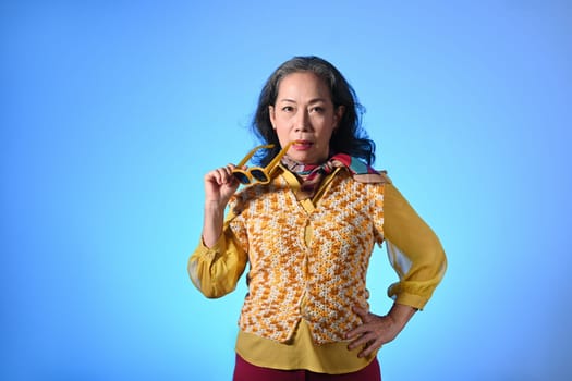 Confident elderly woman with fashionable clothes and sunglasses standing over blue background.