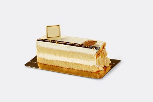 Slice of delectable airy fluffy biscuit cake with delicate light cream decorated with gilded caramelized almond and flakes, presented on gold cardboard against white backdrop