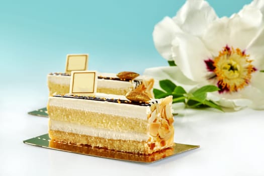 Slices of soft sponge cake with delicate vanilla cream decorated with almond flakes and gilded caramelized nut, presented on pastel blue background with fresh white peony