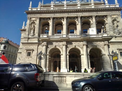 There is a private vehicles parked along the fence, and behind there is a building with columns and statues.