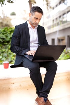 A middle-aged adult man in a suit, drinking coffee, looks at his laptop.