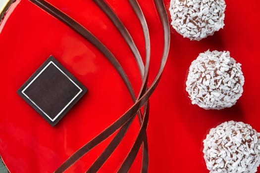 Detail view of glossy red icing covering handmade cake decorated with chocolate curls and coconut truffles, showing texture and design of artisan confectionery