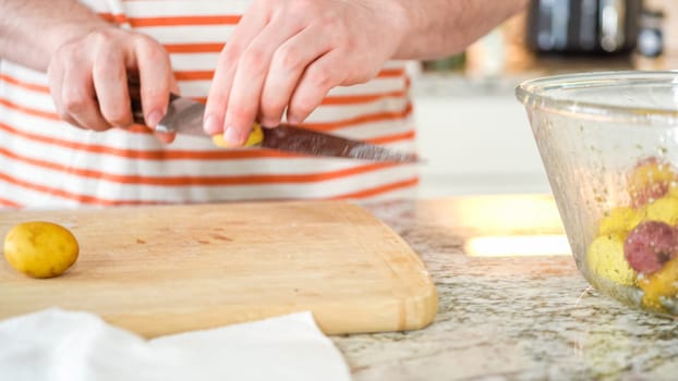 In the contemporary ambiance of a modern kitchen, a young man engages in dinner preparations. His current activity entails meticulously slicing small rainbow potatoes in half on a wooden cutting board, a step towards a well-crafted meal.