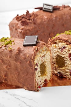 Close-up of sliced chocolate covered loaf cake with soft texture, fruit jelly filling and dried cranberries, adorned with pistachio bits and branded chocolate plaque on marble surface