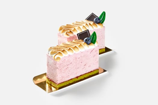 Two airy pink mousse desserts on soft pistachio sponge cake with layer of berry jam, topped with golden toasted meringue, adorned with blueberries and chocolate green leaves, isolated on white