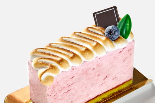 Delectable airy berry mousse cake topped with toasted meringue, accented with fresh blueberry and chocolate decoration, presented on gold cardboard against white backdrop