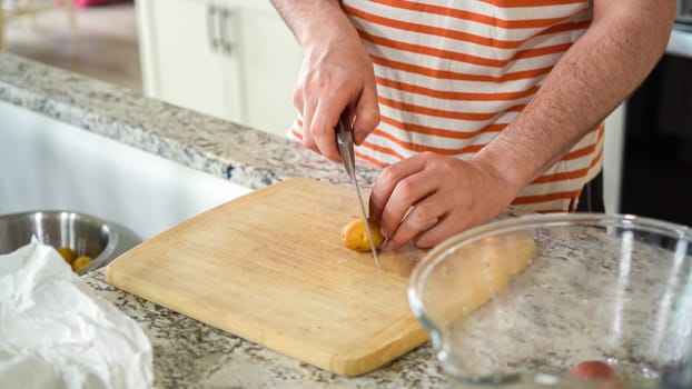 In the contemporary ambiance of a modern kitchen, a young man engages in dinner preparations. His current activity entails meticulously slicing small rainbow potatoes in half on a wooden cutting board, a step towards a well-crafted meal.