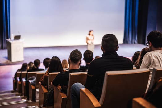 Business and entrepreneurship symposium. Female speaker giving a talk at business meeting. Audience in conference hall. Rear view of unrecognized participant in audience.