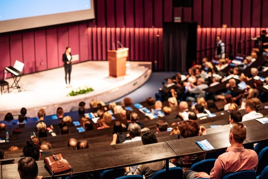 Business and entrepreneurship symposium. Female speaker giving a talk at business meeting. Audience in conference hall. Rear view of unrecognized participant in audience.