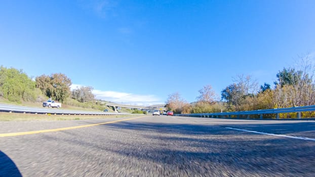 On a crisp winter day, a car cruises along the iconic Highway 1 near San Luis Obispo, California. The surrounding landscape is brownish and subdued, with rolling hills and patches of coastal vegetation flanking the winding road.