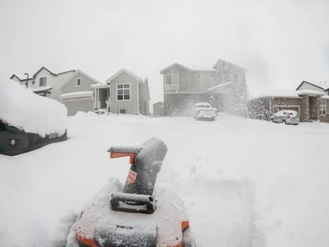 Denver, Colorado, USA-February 4, 2024-View from a snow-blanketed driveway captures a residential area mid-snowfall, with houses partially obscured by the cascading white flakes and a snow-covered car in the foreground.