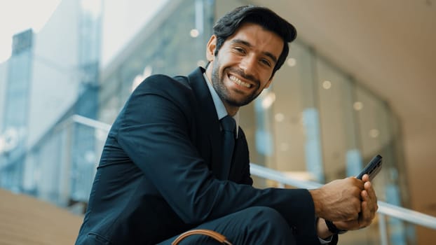 Smiling skilled businessman looking at camera while sitting on stairs. Young professional project manager smile at camera while holding mobile phone at outdoor with blurred background. Exultant.