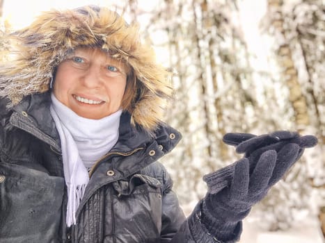 A cheerful middle aged woman in a winter coat with fur, scarf taking selfie on nature outdoors and cold, snowy background with blue sky and white clouds