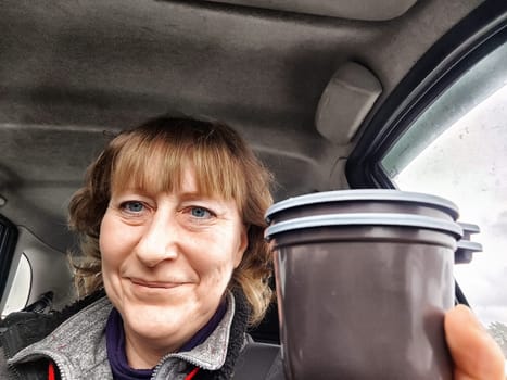 Funny Middle aged woman with a coffee mug in car taking selfie while enjoying drive. Female mature driver posing inside car. Funny happy tourist girl