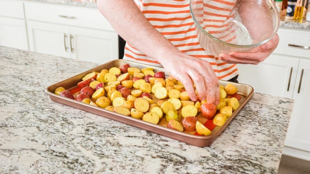 In a modern, white kitchen, a young man is engrossed in dinner preparations. His current endeavor includes arranging seasoned rainbow potatoes on a baking sheet, a meticulous step towards a promisingly tasty meal.