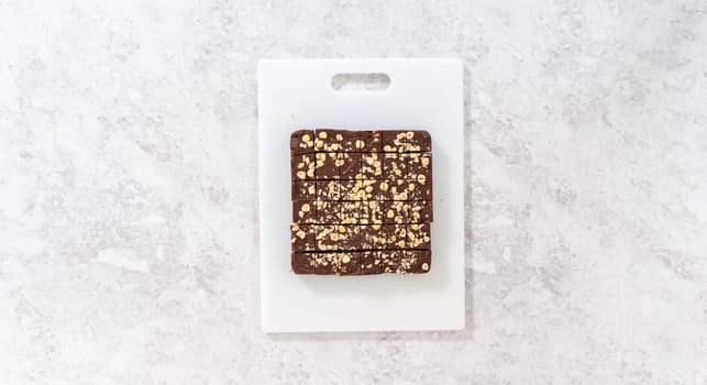 Flat lay. Cutting chocolate hazelnut fudge with a large kitchen knife into square pieces on a white cutting board.