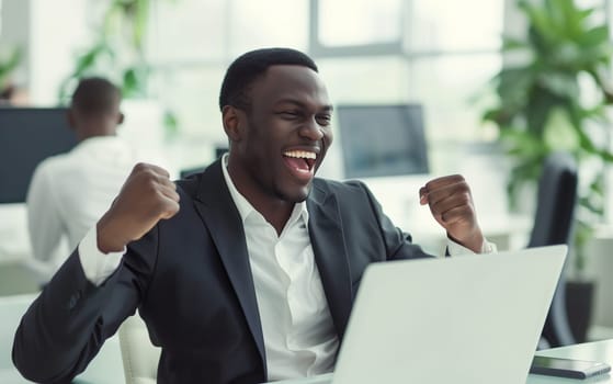 Happy overjoyed laughing african businessman celebrating success raising his hands up while working on laptop sitting at his desk in the office