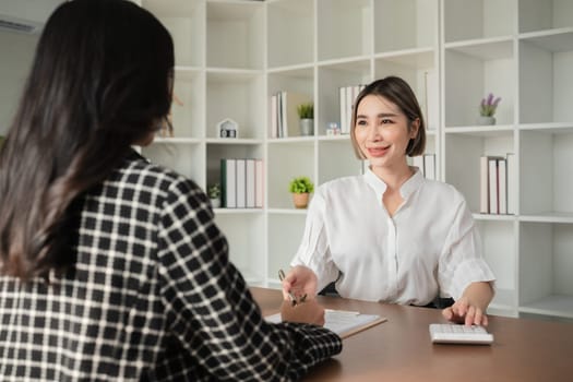 Two Asian businesswomen discussing financial documents with calculator at office desk. Concept of business finance and professional consultation.