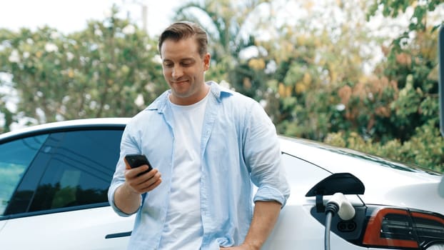 Man use smartphone to pay for electricity at public EV car charging station in nature. Modern environmental and sustainable automobile transportation lifestyle with EV vehicle. Synchronos