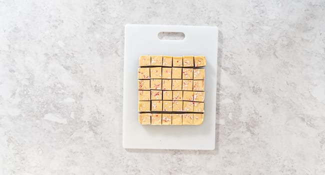 Flat lay. Cutting candy cane fudge with a large kitchen knife into square pieces on a white cutting board.