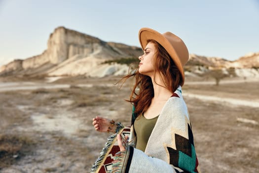 Serene woman in hat standing in field with mountains in background, wrapped in blanket