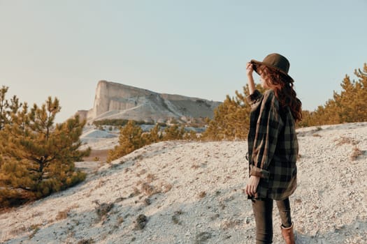 Woman in plaid shirt and hat standing on hill, admiring mountain view on sunny day