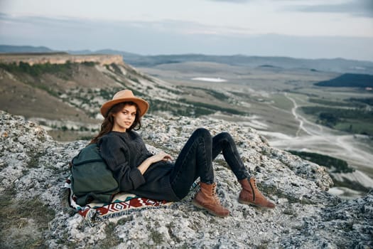 Serene woman in hat sitting with backpack on mountain peak overlooking breathtaking view