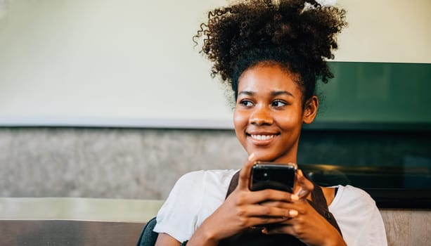 Close-up portrait of a black barista and coffee shop owner using a mobile phone. Smiling woman in uniform working communicating joyfully. Entrepreneur's relaxation is apparent.
