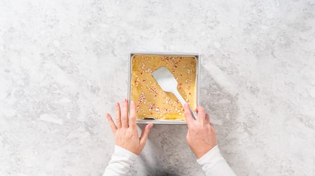 Flat lay. Filling square cheesecake pan lined with parchment paper with fudge mixture to prepare candy cane fudge.