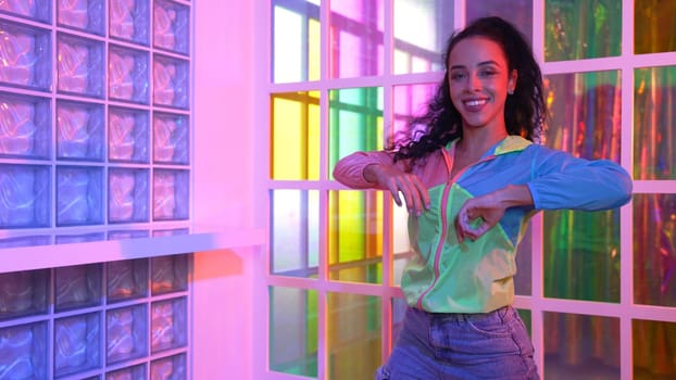 Happy hispanic girl in lively mood moving along music in neon light while wearing colorful cloth at night club. Street dancer making energetic footstep or movement while looking at camera. Regalement.