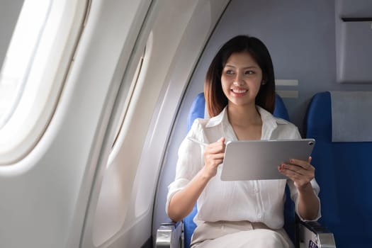 Asian woman using tablet on airplane. Concept of air travel, digital technology, and in-flight entertainment.