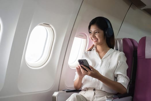 Asian woman using smartphone and headphones on airplane. Concept of air travel, technology, and in-flight entertainment.