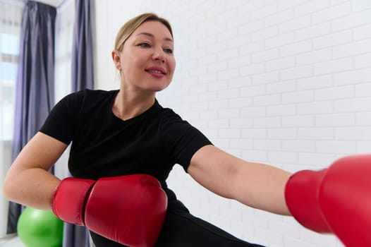 Smiling athlete woman boxer fighter stretching and warming up her body before intensive boxing training, wearing red boxing gloves, exercising over white studio background. People and active lifestyle