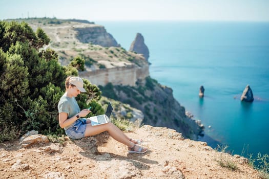 A woman is sitting on a rocky hillside overlooking the ocean. She is using a laptop and she is enjoying the view