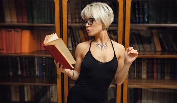 Portrait of an adult beautiful girl with glasses and short hair, with a book in her hand in a room against the background of a library