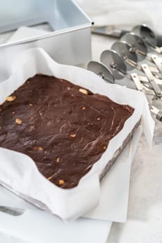 Removing chocolate macadamia fudge from a square cheesecake pan lined with parchment.