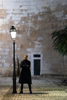 A man stands in front of a street lamp, looking at the camera. The scene is set in a city at night, with a bench nearby. The woman is wearing a black coat and she is waiting for someone or something
