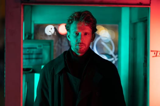 A man is standing in front of a neon sign. The sign is green and red. The man is wearing a black coat and a black hat