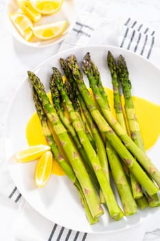 Serving steamed asparagus with homemade hollandaise sauce with lemon wedges on a white plate.