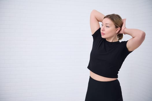 Beautiful Caucasian athletic woman 30s in black sportswear, tying a ponytail, confidently looking way, isolated over white wall background with copy advertising space. People and martial art concept