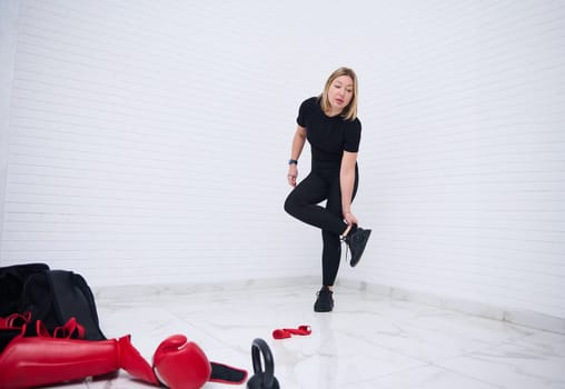 Full size shot of an European young sporty woman 30s, in black sports clothes, standing against white wall background. Sports equipment on the foreground. People, fitness and sport concept