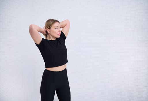 Beautiful Caucasian European athletic woman 30s in black sportswear, tying a ponytail, isolated over white wall background