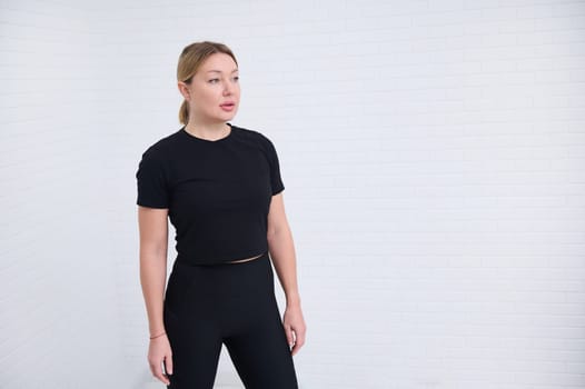 Beautiful European athletic woman 30s in black sportswear, confidently looking way, isolated over white wall background with copy advertising space