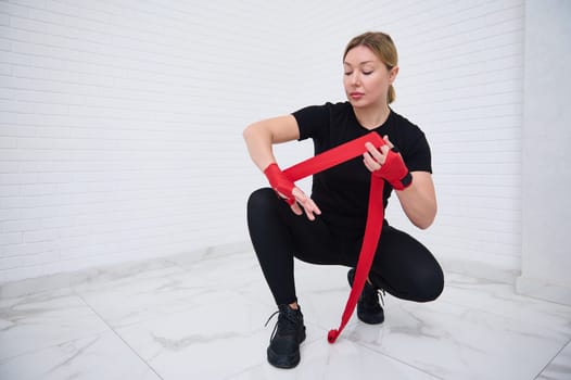 Confident portrait of determined authentic European young woman boxer 35-39 years old in black active wear sitting in squat position, typing tapes over white background, preparing for boxing training