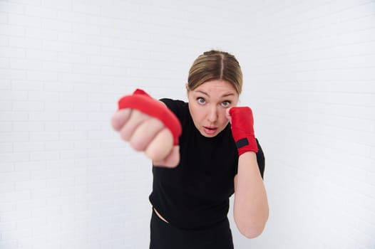 Athletic woman, boxer with red tapes around her fists, punching forward while practicing boxing training over white isolated background. Copy space for text. People, sport and martial art concept