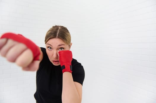 Athletic woman fighter with red tapes around her fists, punching forward while practicing boxing exercises over white isolated background. Copy space for text. People, sport and martial art concept