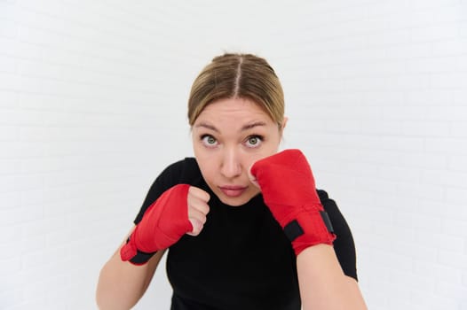 Strong and beautiful European athletic woman, boxer fighter with kickboxing red tapes, doing some boxing exercises, punching forward to camera. Martial art and combat. Wellness, activities and sports